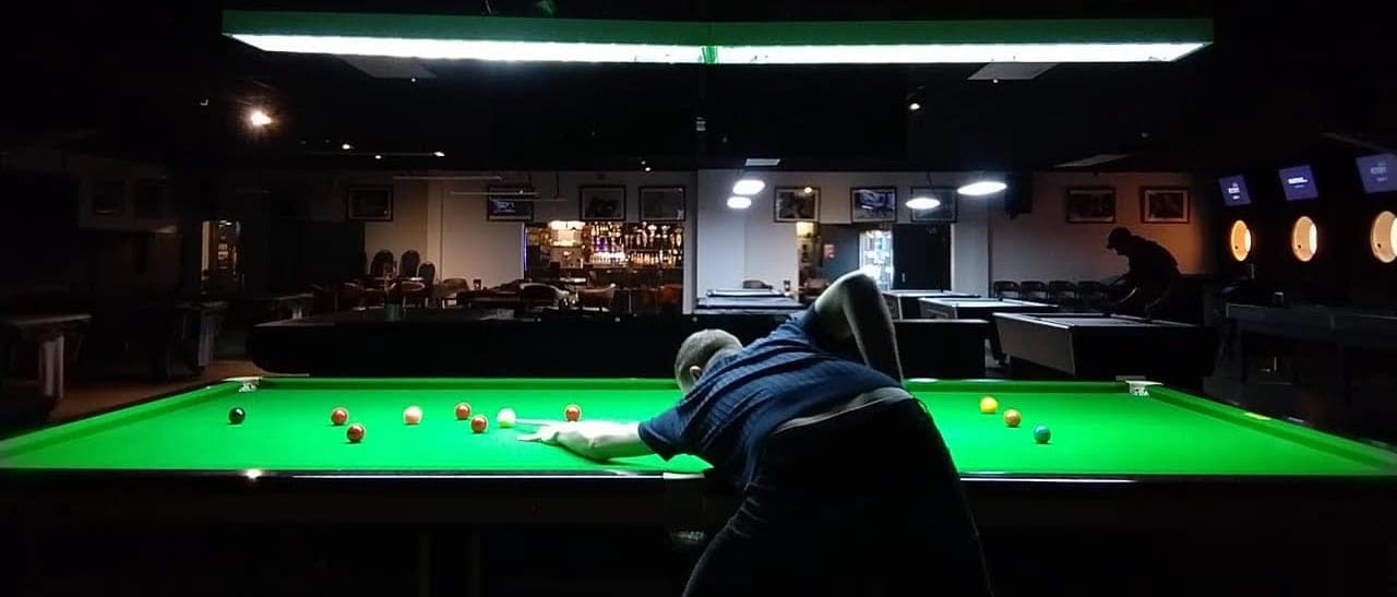 A player down on a shot at a snooker table under the long table light, the club's bar and pool tables behind